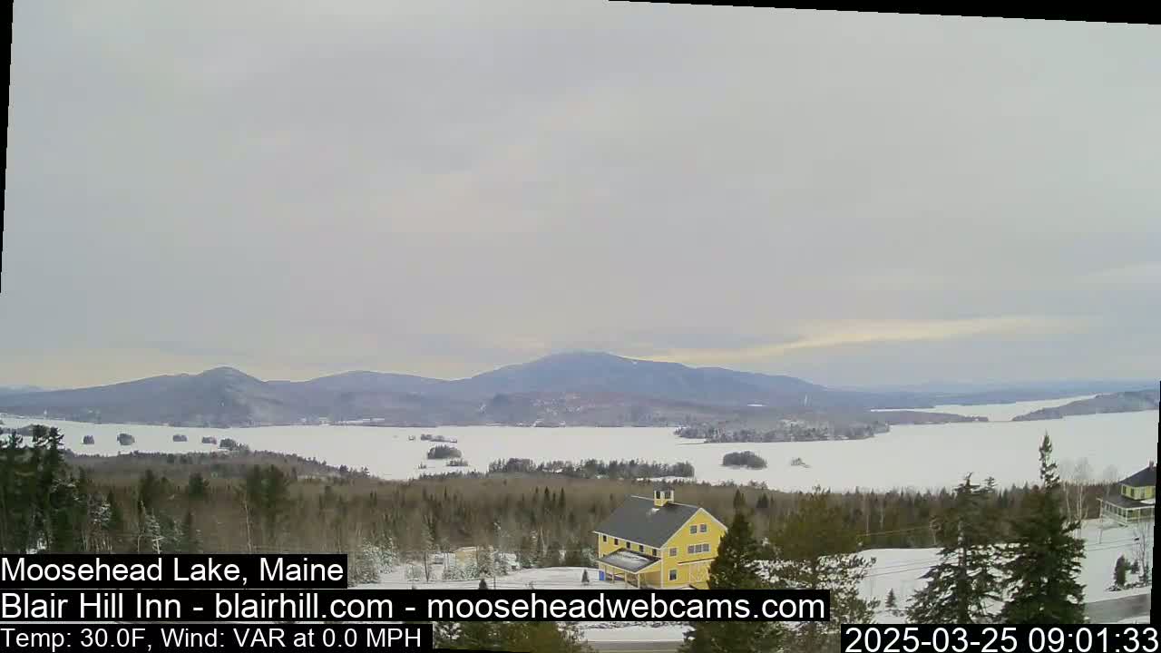 A snow-covered lake stretches to distant mountains under an overcast sky, with a yellow building and evergreen trees in the foreground.