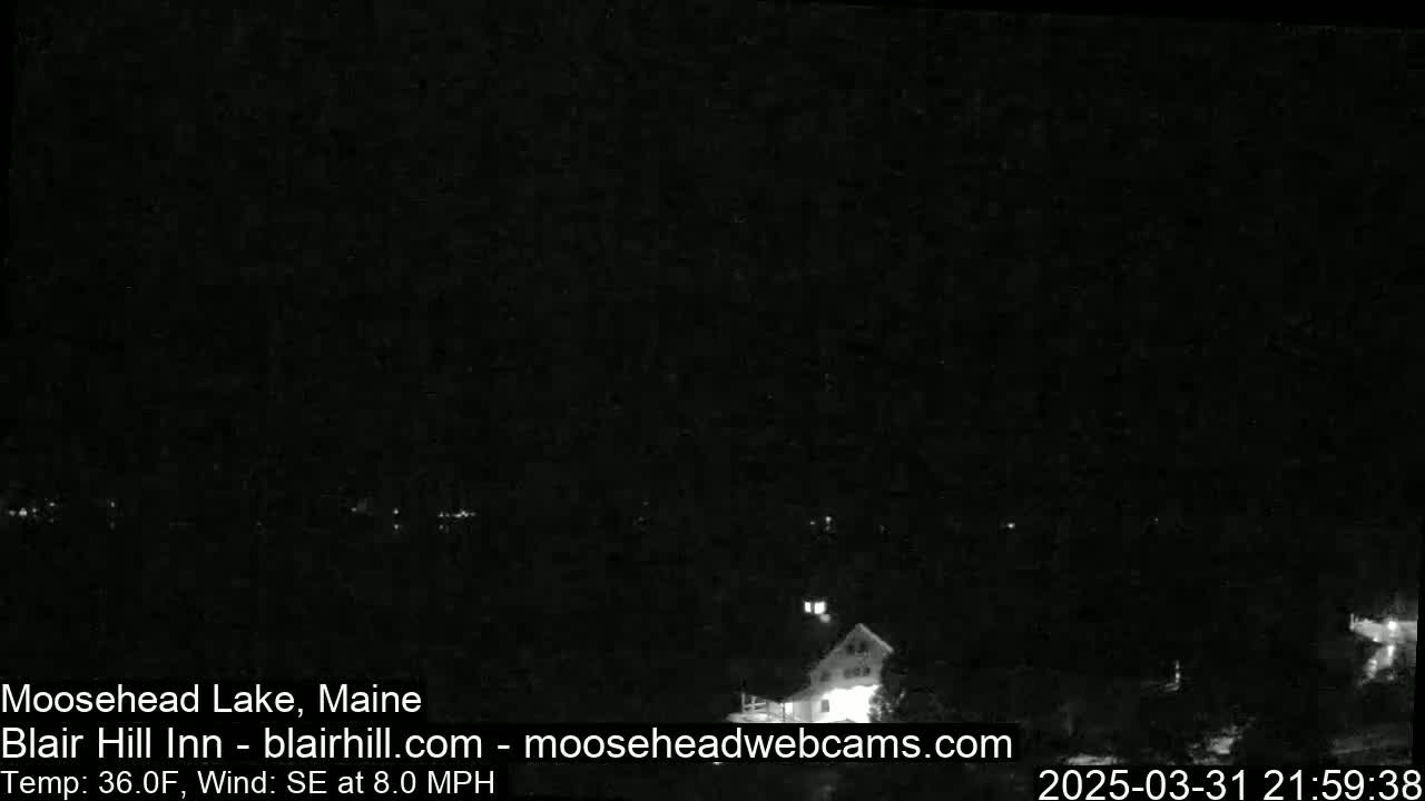 A nighttime, low-light view of a dark landscape with a few faint lights and a building visible in the distance, under a dark sky with a southeasterly wind at 8 mph and a temperature of 36°F.