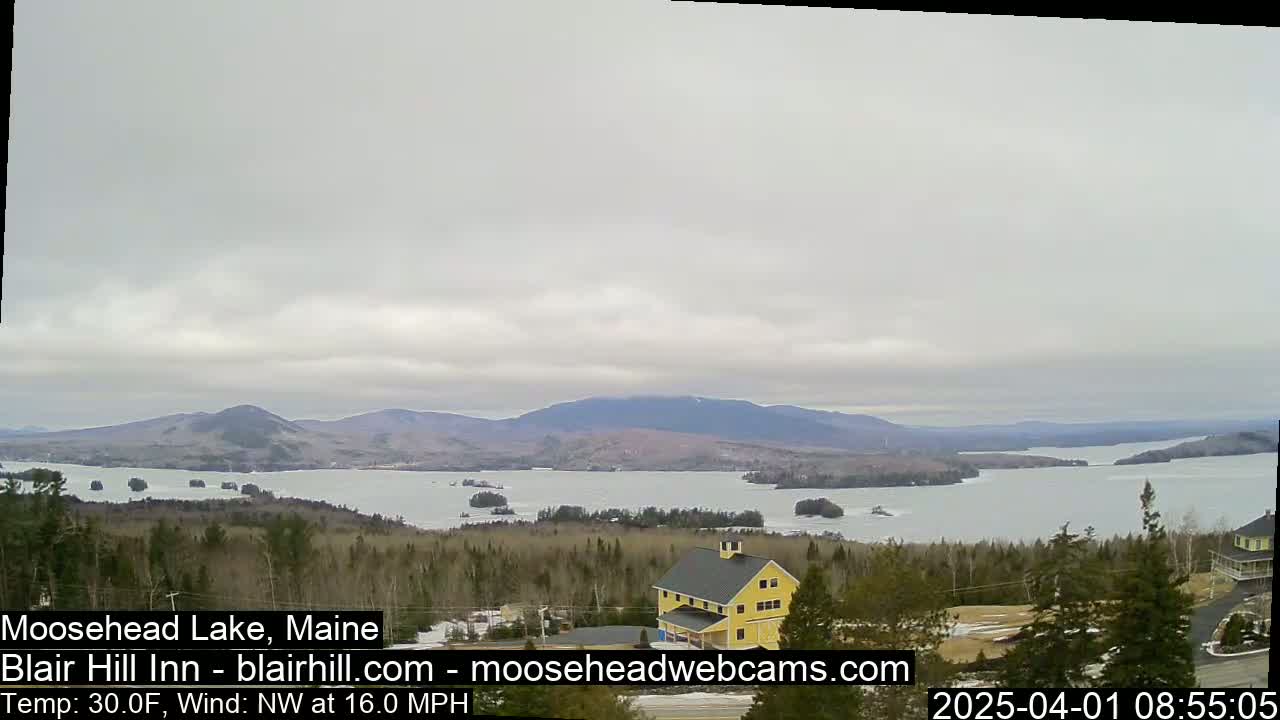 A mostly cloudy day reveals a view of Moosehead Lake, partially frozen, surrounded by dark green trees and distant mountains, with a yellow building visible in the foreground.