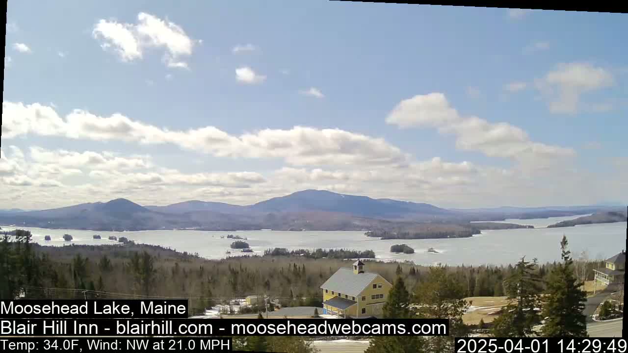 A partly cloudy day reveals a view of a lake partially covered in ice, surrounded by a wooded landscape and distant mountains.