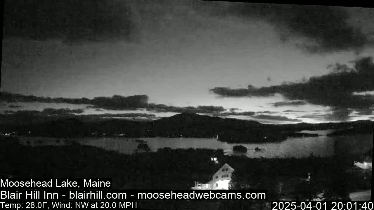 A nighttime view of a lake and mountains under a cloudy sky with a northwest wind at 20 mph and a temperature of 28°F.