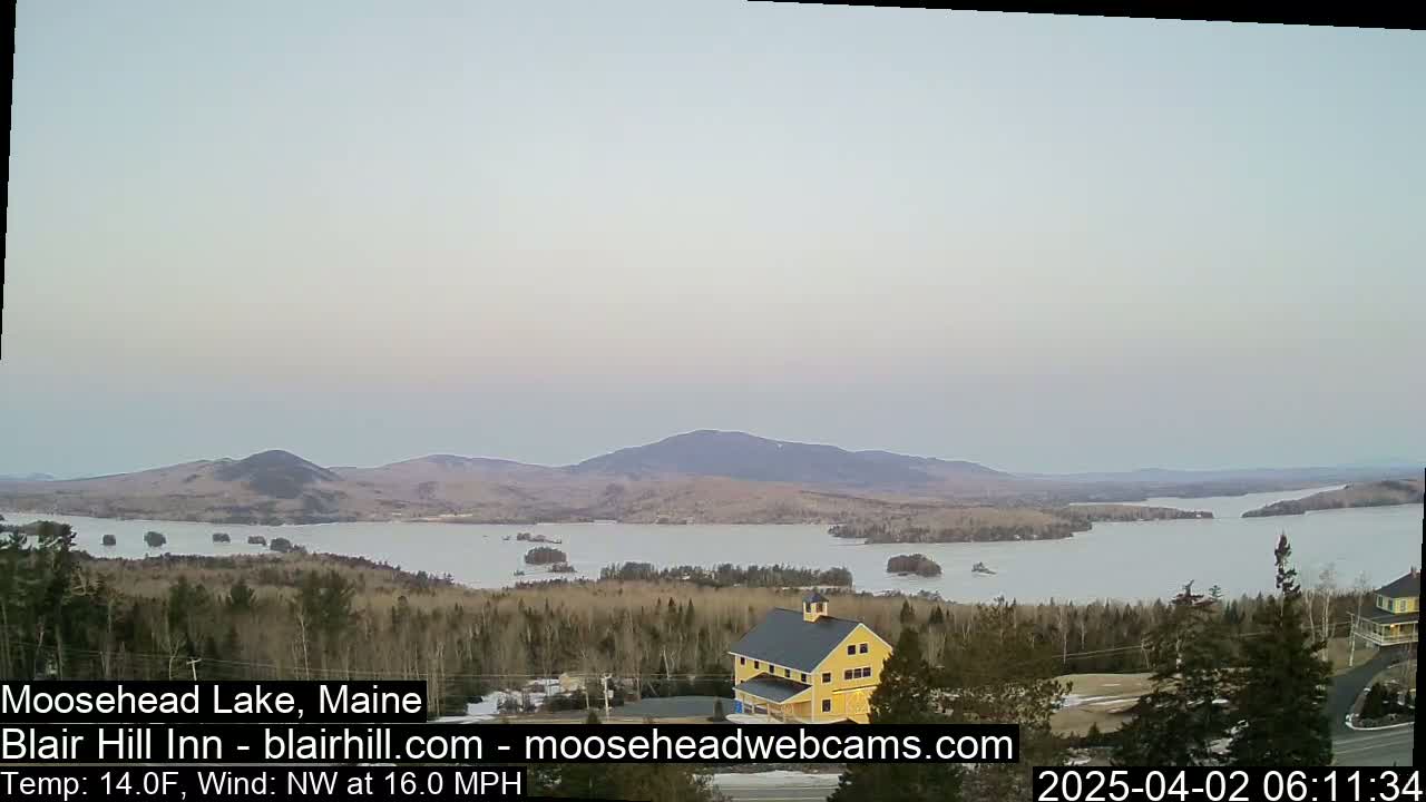 A partly frozen lake and distant mountains are visible under a pale sky, with a building situated in the foreground amidst a sparsely wooded area.