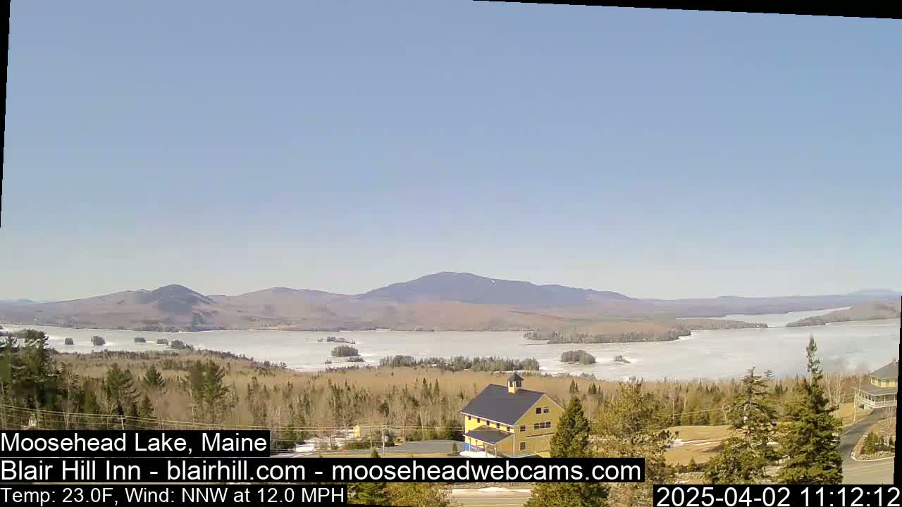 A partially frozen lake stretches to distant mountains under a clear blue sky, with a yellow building and evergreens in the foreground.