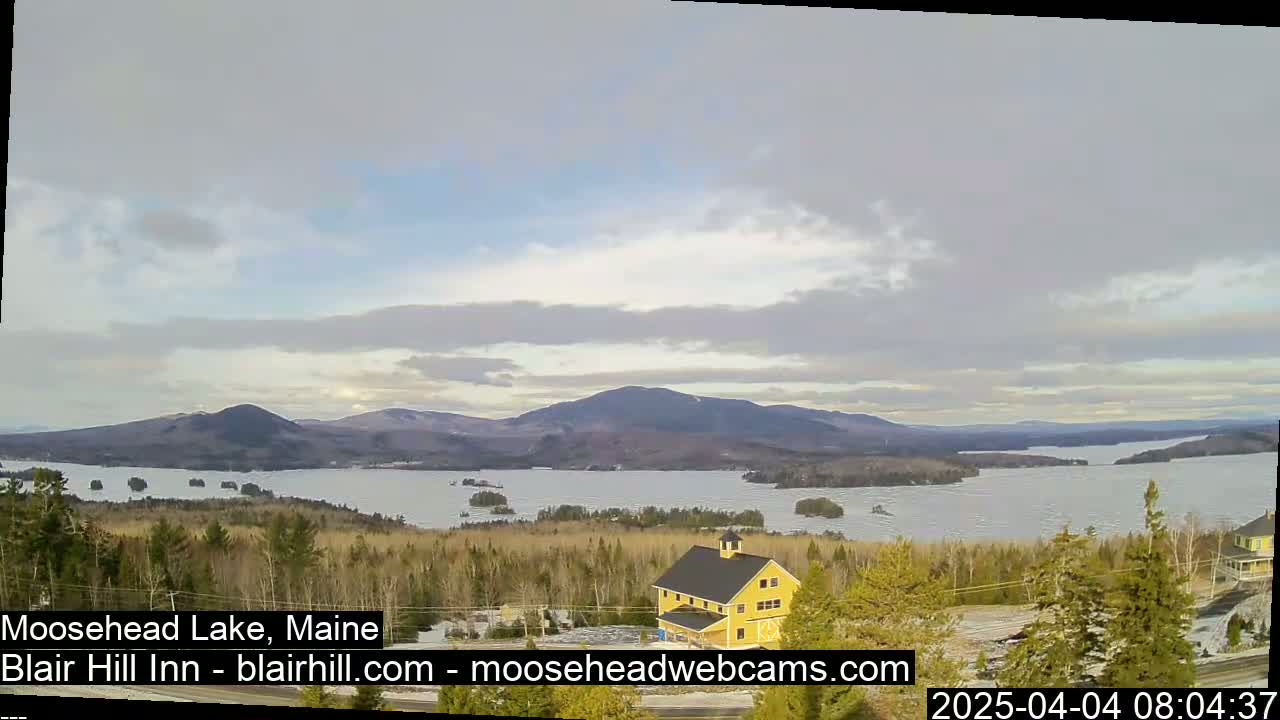 A mostly cloudy day reveals a view of a large lake partially covered in ice, surrounded by snow-dusted trees and distant mountains.
