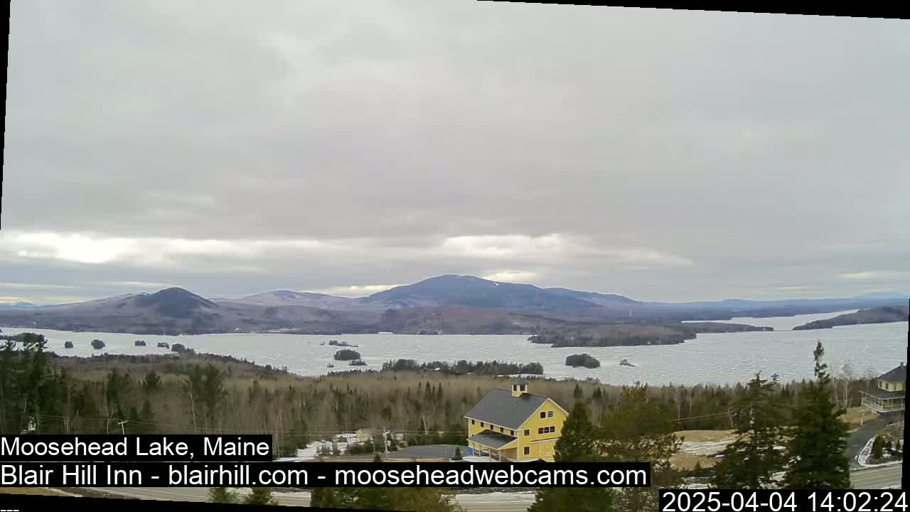 A partly cloudy day reveals a wide, partially frozen lake nestled between low, snow-dusted mountains, with a yellow building visible in the foreground.
