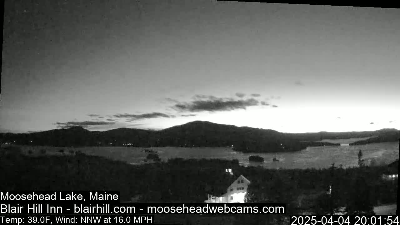 A grayscale image shows a calm lake at dusk, with mountains in the background and a building in the foreground, under a partly cloudy sky with a 16 mph north-northwest wind and a temperature of 39°F.