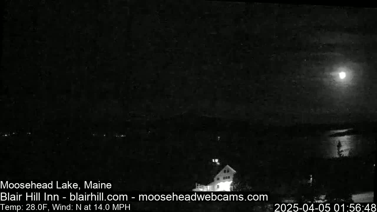 A nighttime long shot of a lake and a building under a bright moon, with a north wind at 14 mph and a temperature of 28 degrees Fahrenheit.