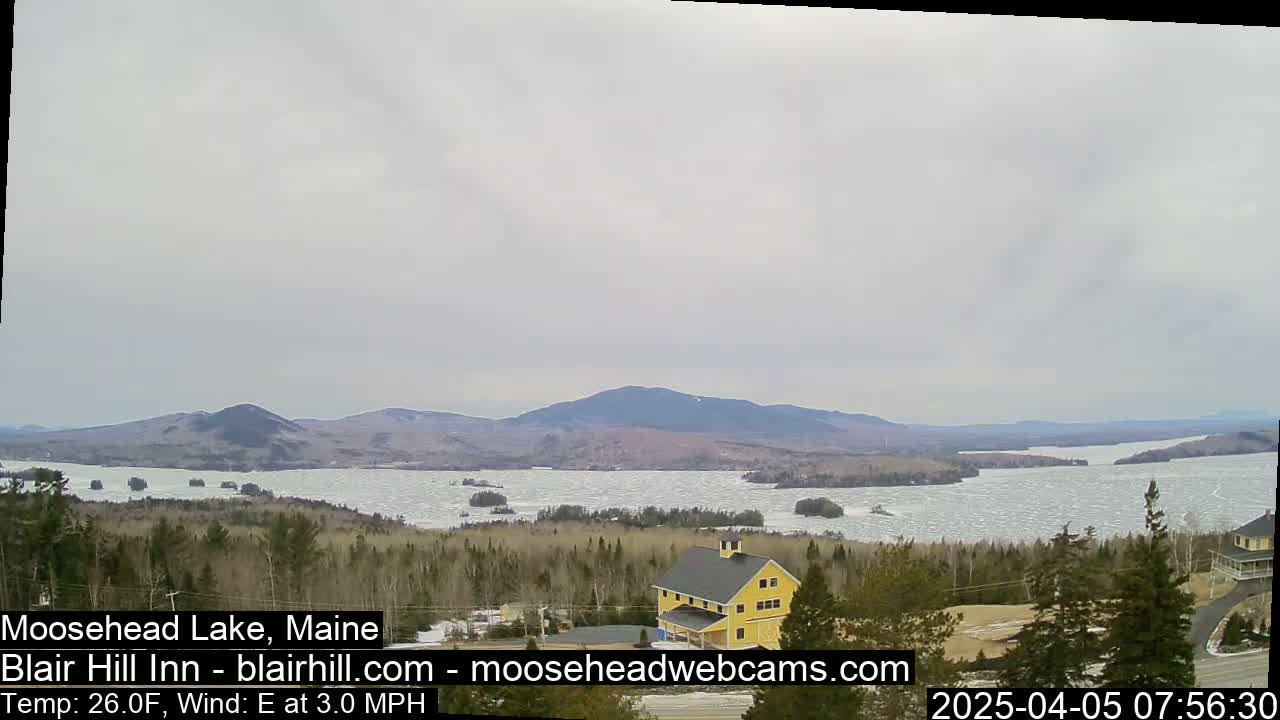 A partially ice-covered lake stretches towards distant mountains under an overcast sky, with a yellow building visible in the foreground.