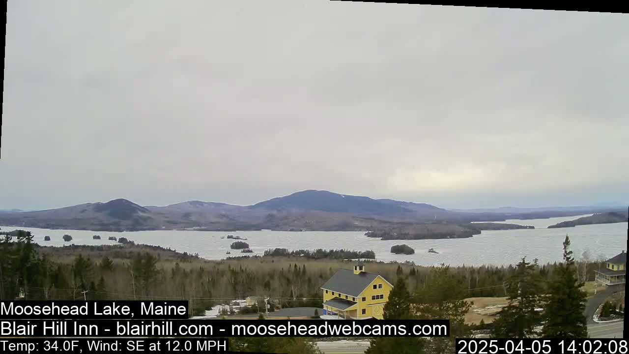 A mostly cloudy view of a large lake partially covered in ice, with distant mountains and a yellow building in the foreground.