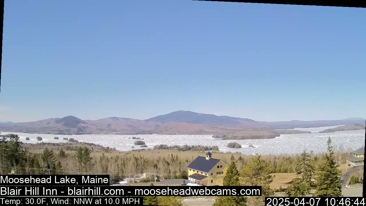 A partly frozen lake stretches to distant mountains under a clear blue sky, with a yellow building visible in the foreground.