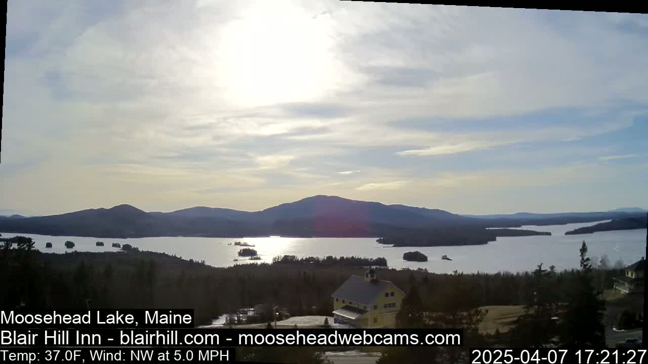 A large lake surrounded by mountains and trees is seen under a partly cloudy sky, with a building visible in the foreground.