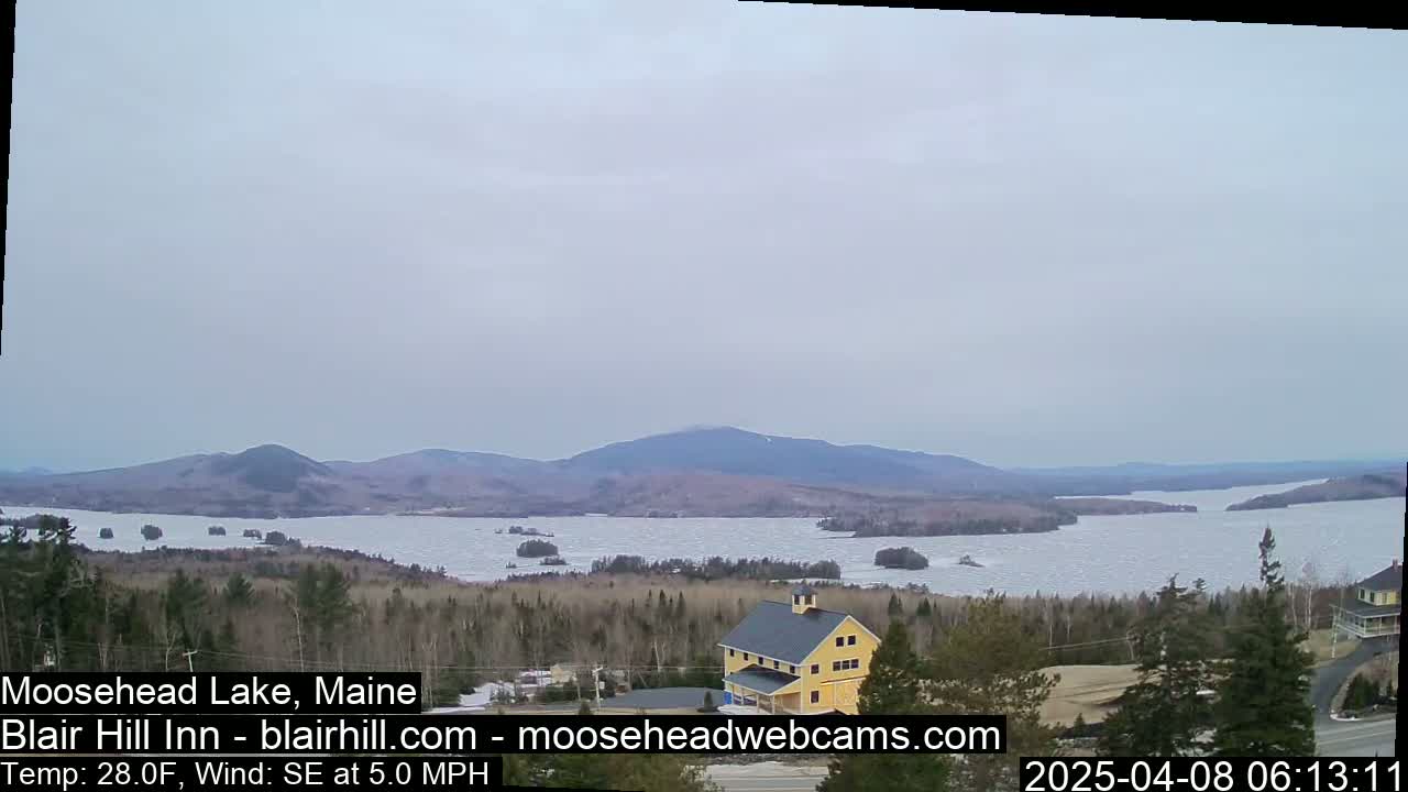 A partly frozen lake stretches towards distant mountains under an overcast sky, with a yellow building visible in the foreground.