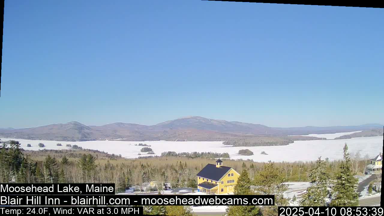 A partially frozen lake is seen from a high vantage point on a sunny day, with a yellow building and snow-covered landscape in the foreground and mountains in the background.