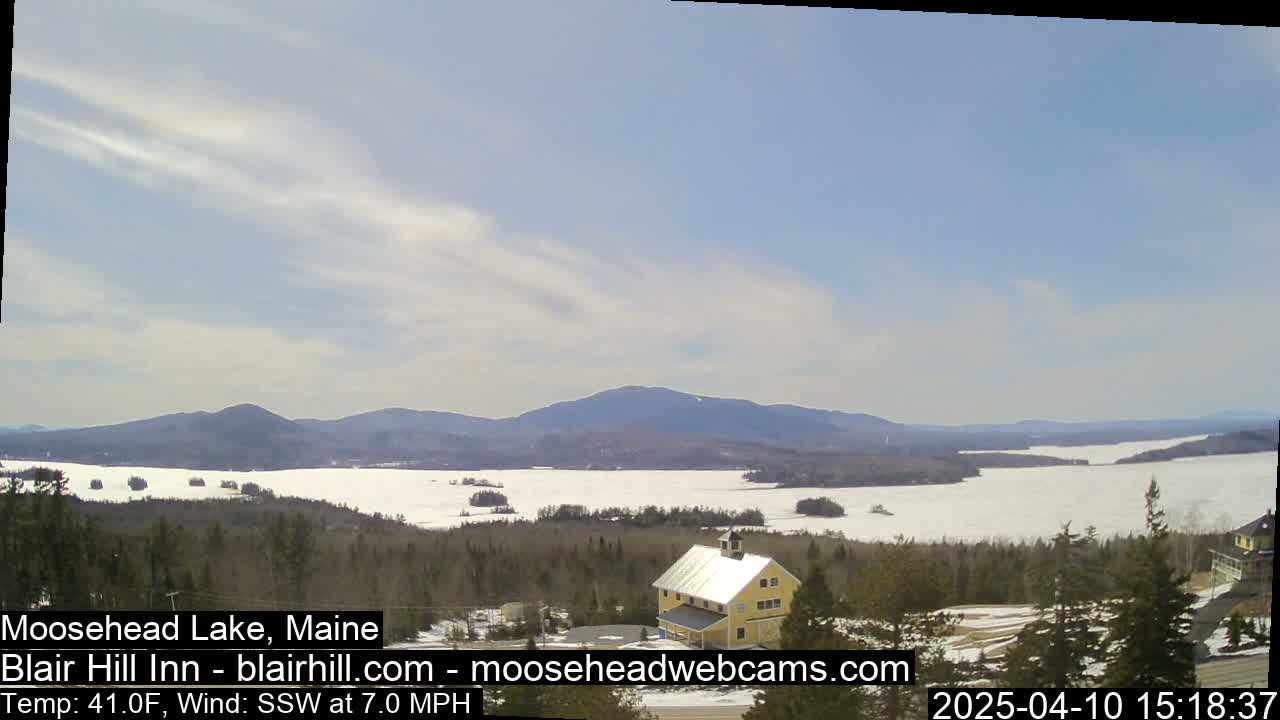 A partly cloudy day reveals a partially frozen lake nestled among snow-dusted trees and distant mountains.