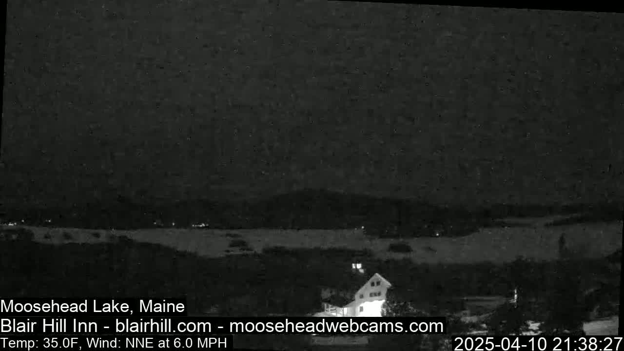 A nighttime view of a snow-covered landscape with a lake and a building, under a dark sky with a light wind.