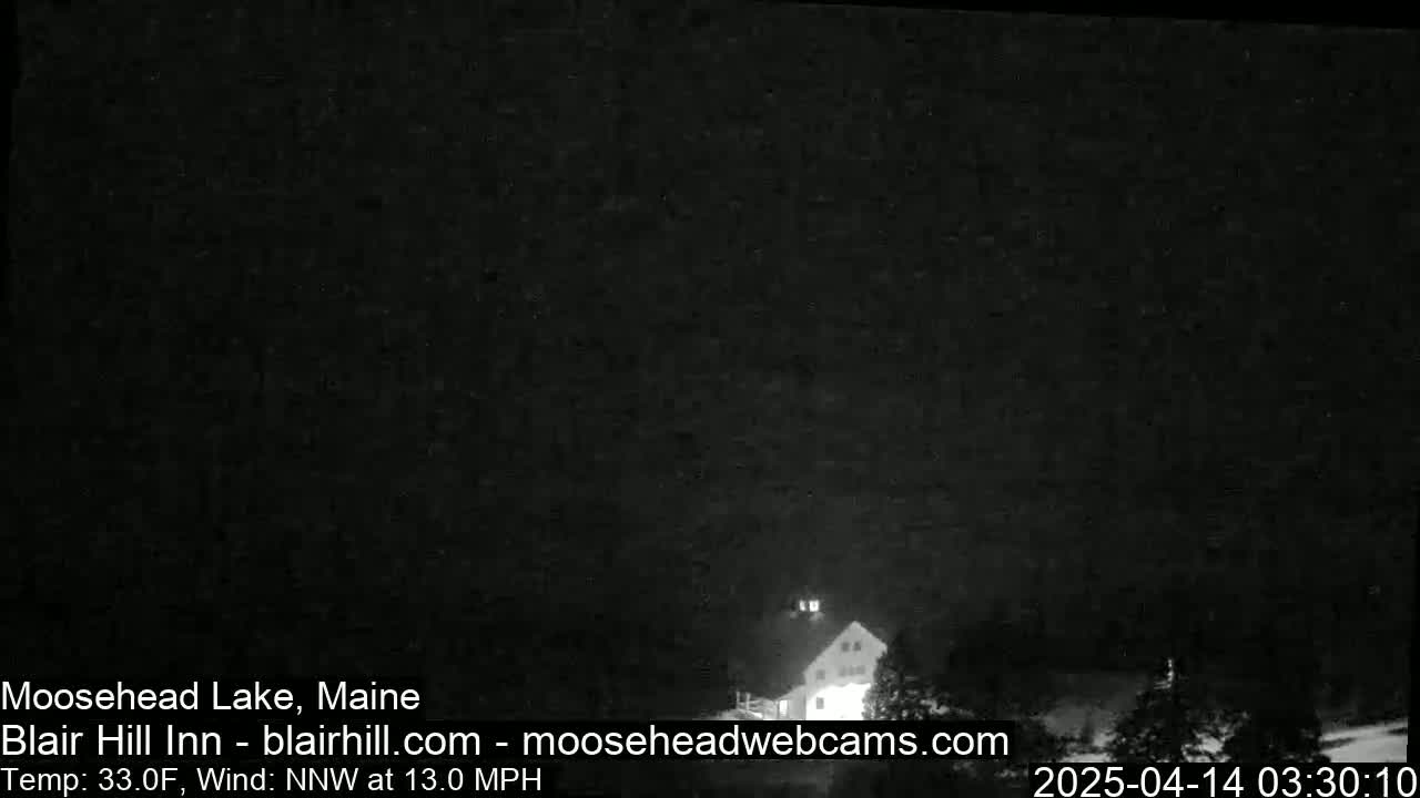 A dark, nighttime image shows a dimly lit building amidst trees under a cloudy sky, with a 33°F temperature and 13 mph northwest wind.
