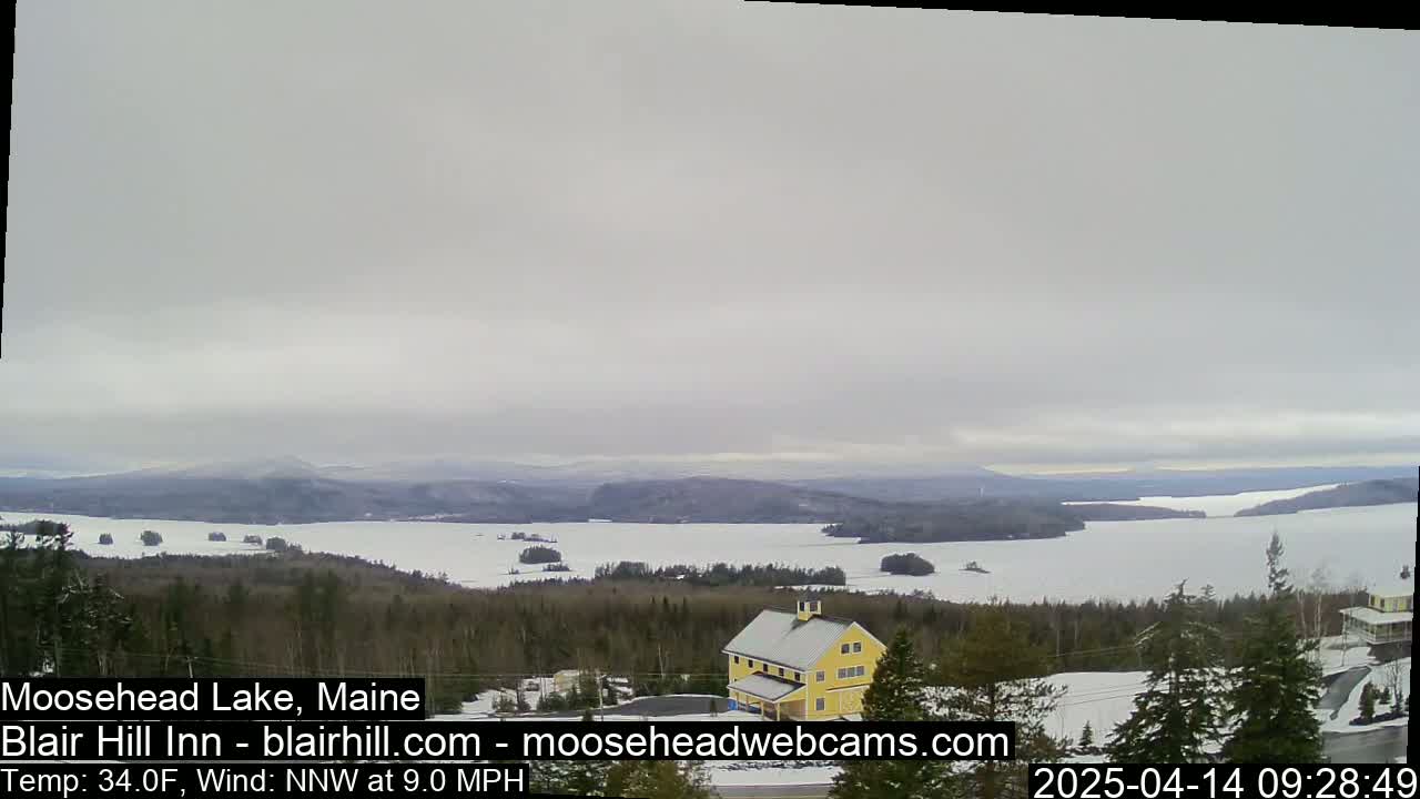 A mostly cloudy day reveals a view of a large snow-covered lake, surrounded by evergreen trees and a yellow building, with distant mountains visible in the background.