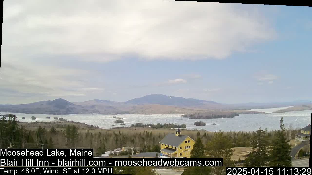 A partly cloudy day reveals a partially frozen lake nestled among hills and mountains, with a yellow building situated in the foreground.