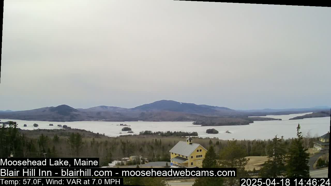 A partly frozen lake is visible from a high vantage point, under an overcast sky, with a yellow building and dark green trees in the foreground, and mountains in the background.