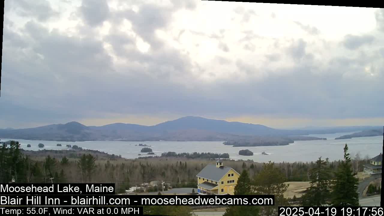 A mostly cloudy sky overlooks a large lake surrounded by trees and mountains, with a yellow building visible in the foreground.