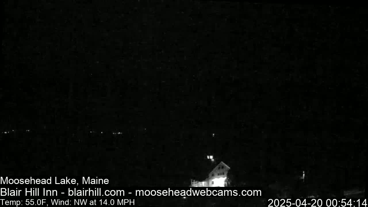 A nighttime, low-light view of a lake and a building with lights on, under a clear, dark sky with a northwest wind at 14 mph and a temperature of 55°F.