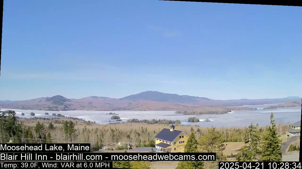 A yellow building sits on a hill overlooking a partially frozen lake and distant mountains under a clear blue sky.