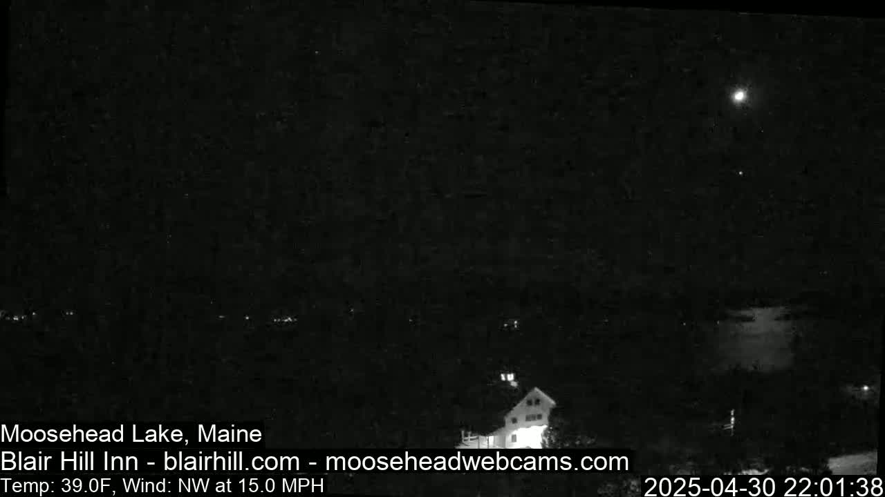 A nighttime, low-light image shows a dark landscape under a partly cloudy sky with a bright light source possibly from the moon, with a northwest wind at 15 mph and a temperature of 39°F.