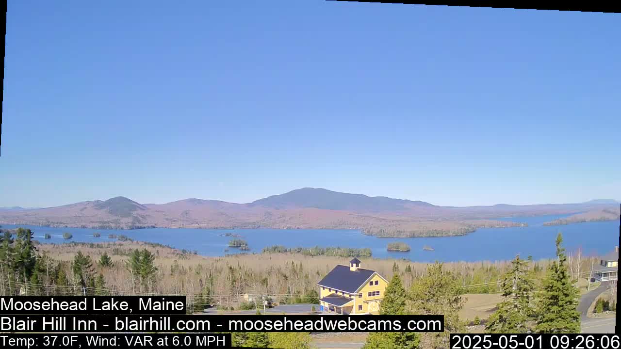 A large lake is visible, surrounded by trees and mountains under a clear blue sky, with a yellow building in the foreground.