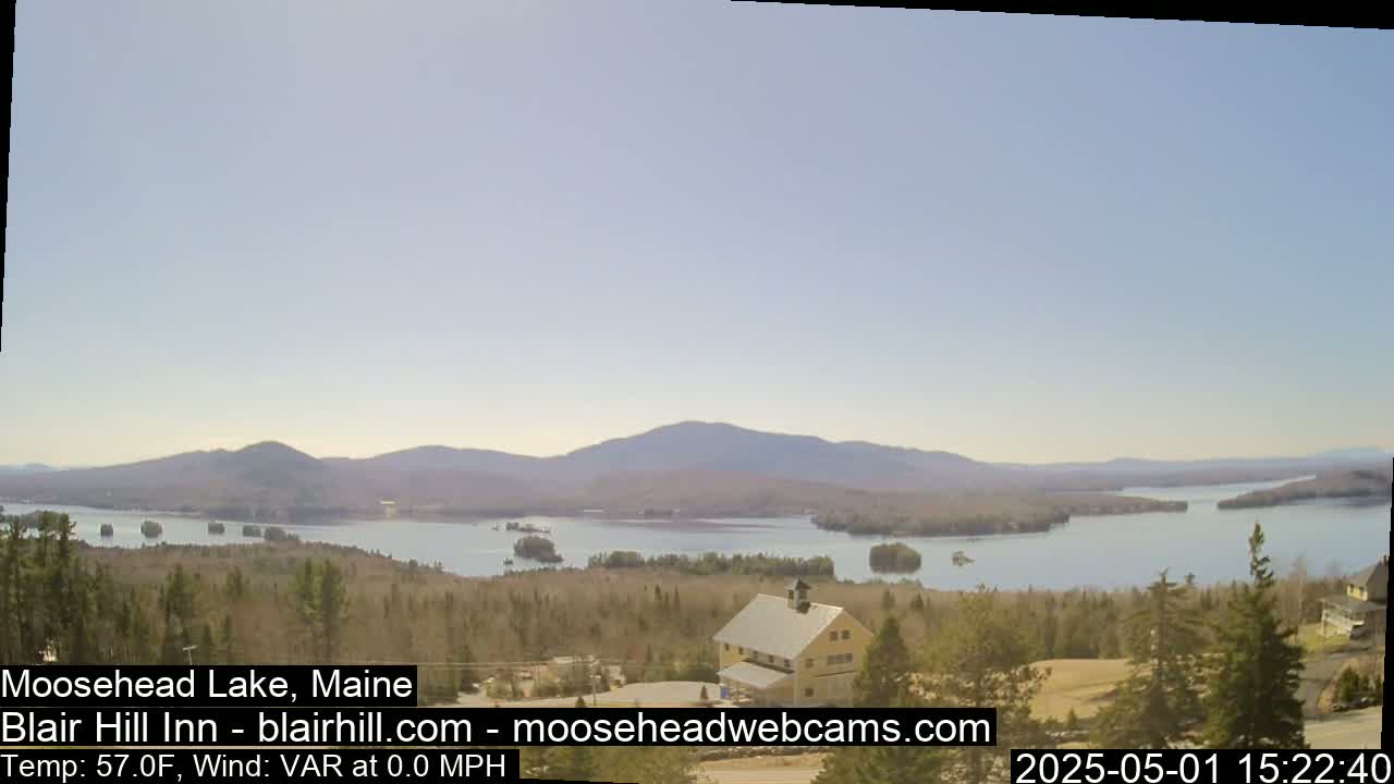 A large lake with many islands is seen from an elevated vantage point on a clear, calm day, with a building and trees in the foreground and mountains in the background.