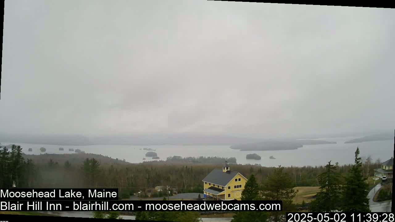 A large lake is visible under an overcast sky, with a yellow building and evergreen trees in the foreground.