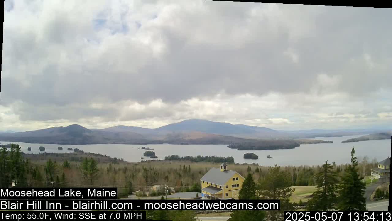 A partly cloudy day reveals a view of a large lake surrounded by mountains and evergreen trees, with a yellow building visible in the foreground.