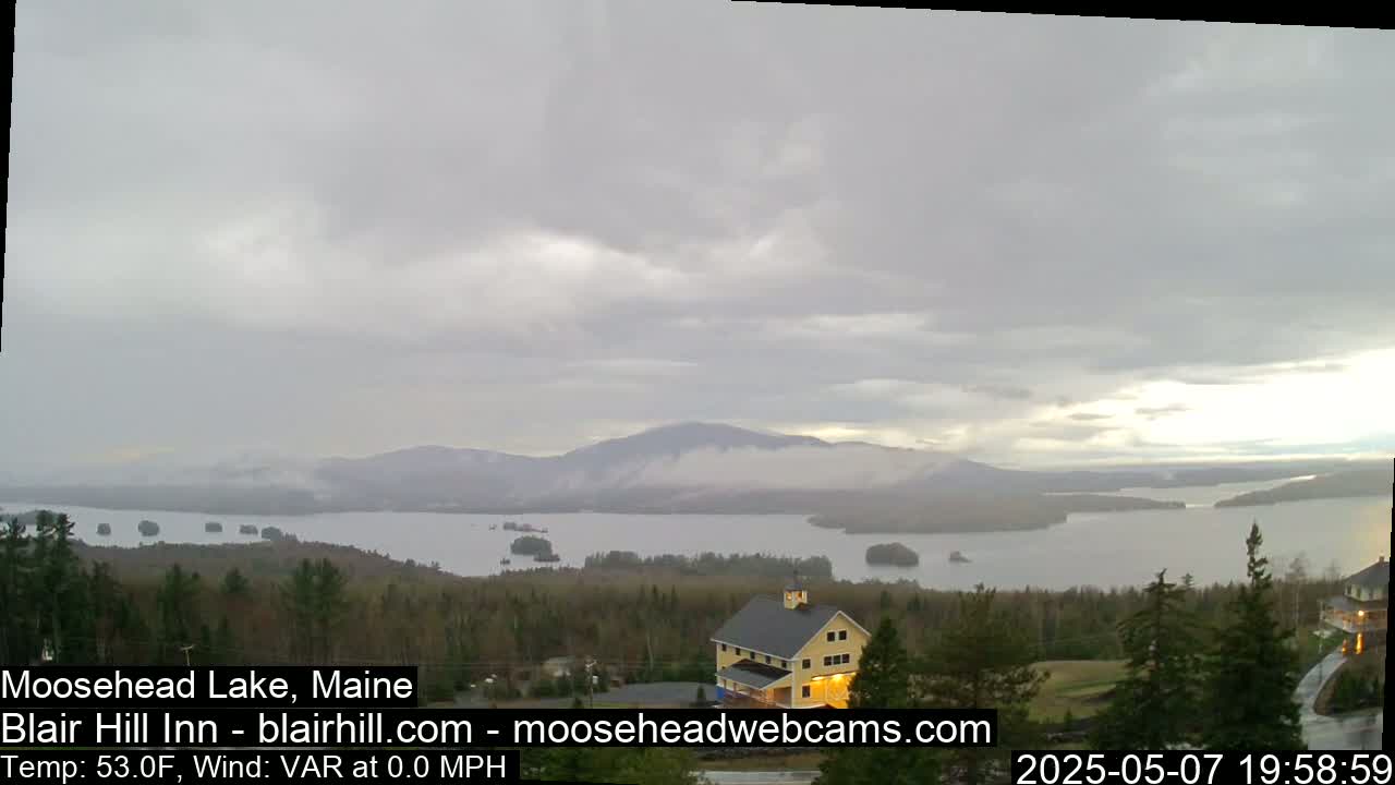 A large lake surrounded by mountains and trees is viewed from a high vantage point under an overcast sky, with a building visible in the foreground.