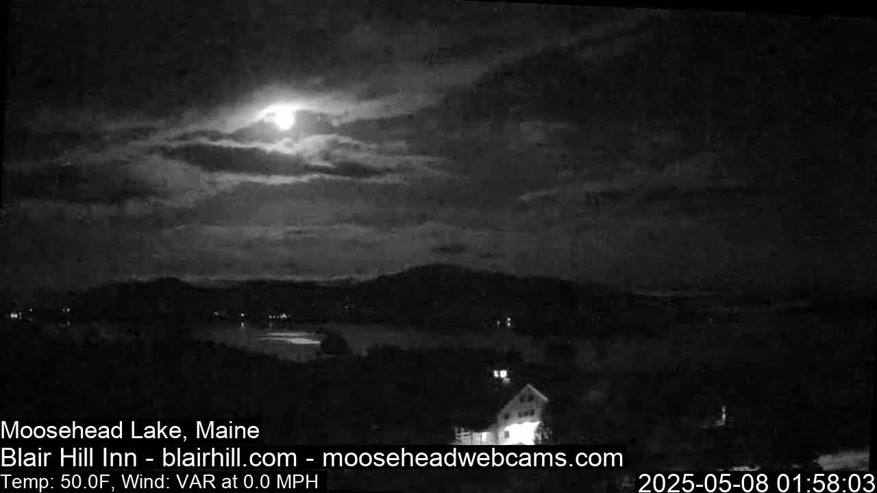 A nighttime view of a lake and mountains under a partly cloudy sky with a bright moon.