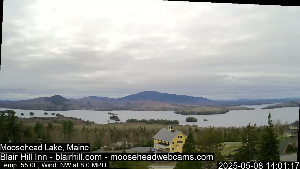 A wide shot shows a calm lake surrounded by forested hills and mountains under an overcast sky.