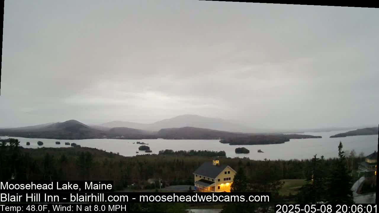 A large lake is seen from an elevated vantage point, under an overcast sky, with a yellow building and dark green trees in the foreground, and mountains in the distance.