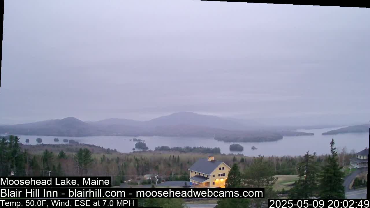 A hazy, calm evening view of a lake surrounded by mountains and trees, with a yellow building visible in the foreground.
