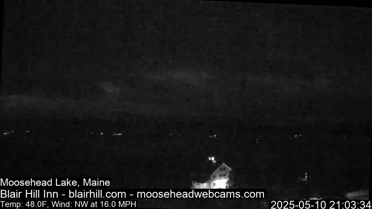 A nighttime long shot of a lake and a building under a dark, cloudy sky with a northwest wind of 16 mph and a temperature of 48°F.