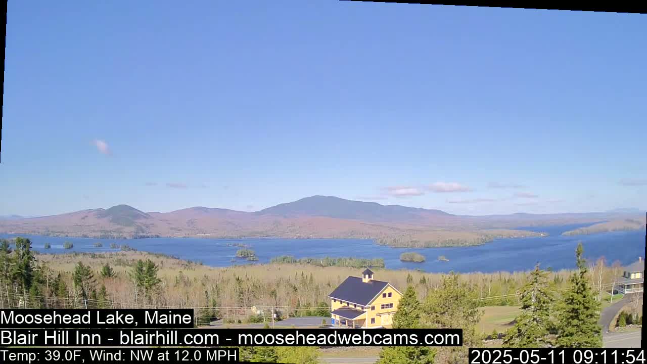 A large lake surrounded by trees and mountains is visible under a clear blue sky with a few clouds, a yellow building is in the foreground.