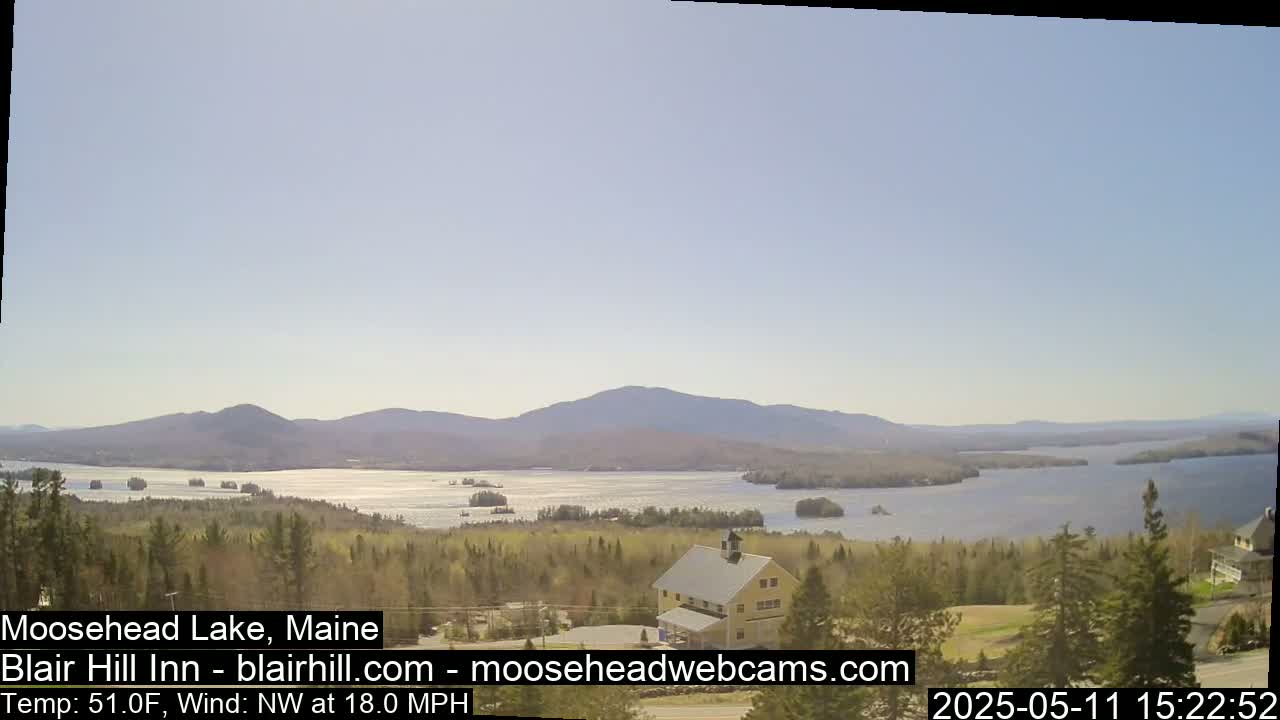 A large lake surrounded by mountains and trees is visible from a high vantage point, under a mostly clear sky with a northwest wind.