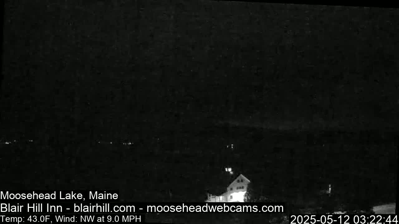 A nighttime, low-light image shows a dark landscape with a building faintly visible in the lower right, under a mostly clear sky with a northwest wind.
