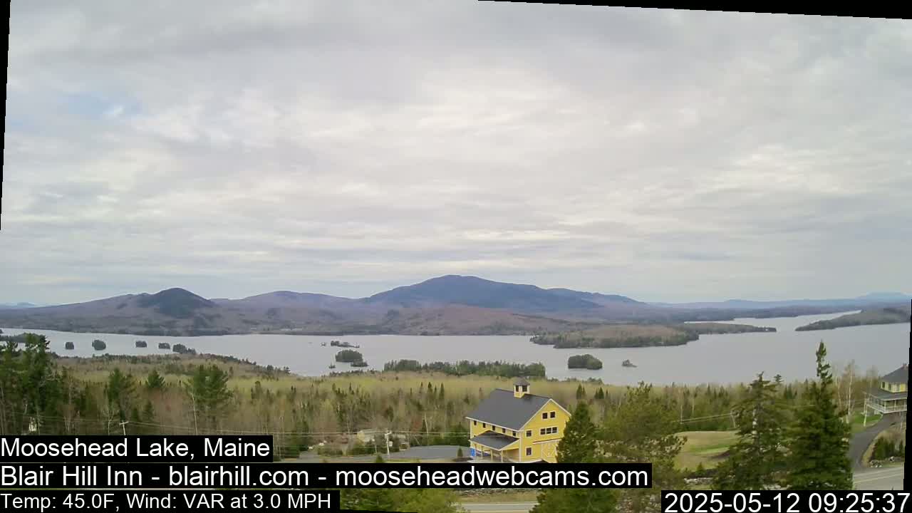 A yellow building sits on a hill overlooking a calm lake surrounded by spring green trees and mountains under a cloudy sky.