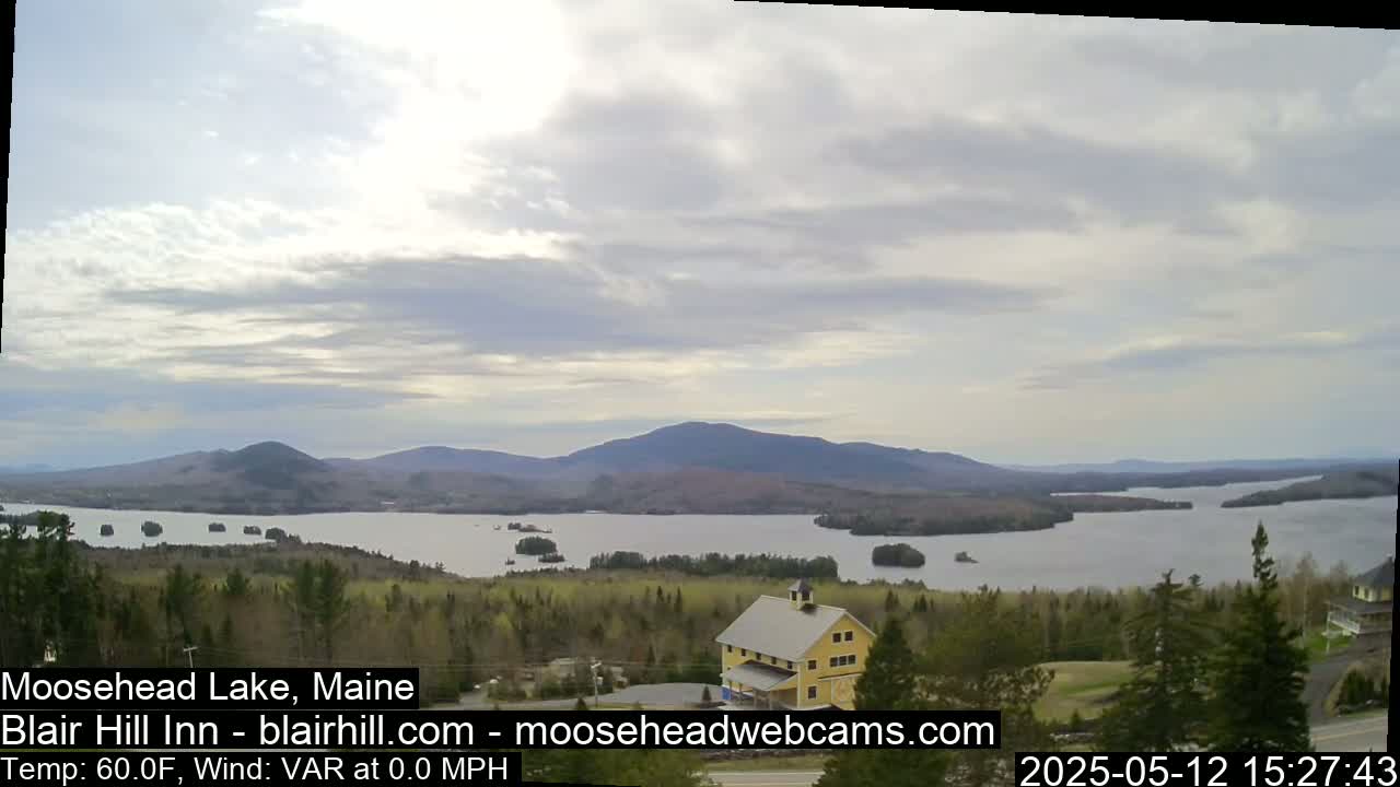 A large lake surrounded by forested hills and mountains under a partly cloudy sky is seen from an elevated vantage point, with a yellow building visible in the foreground.