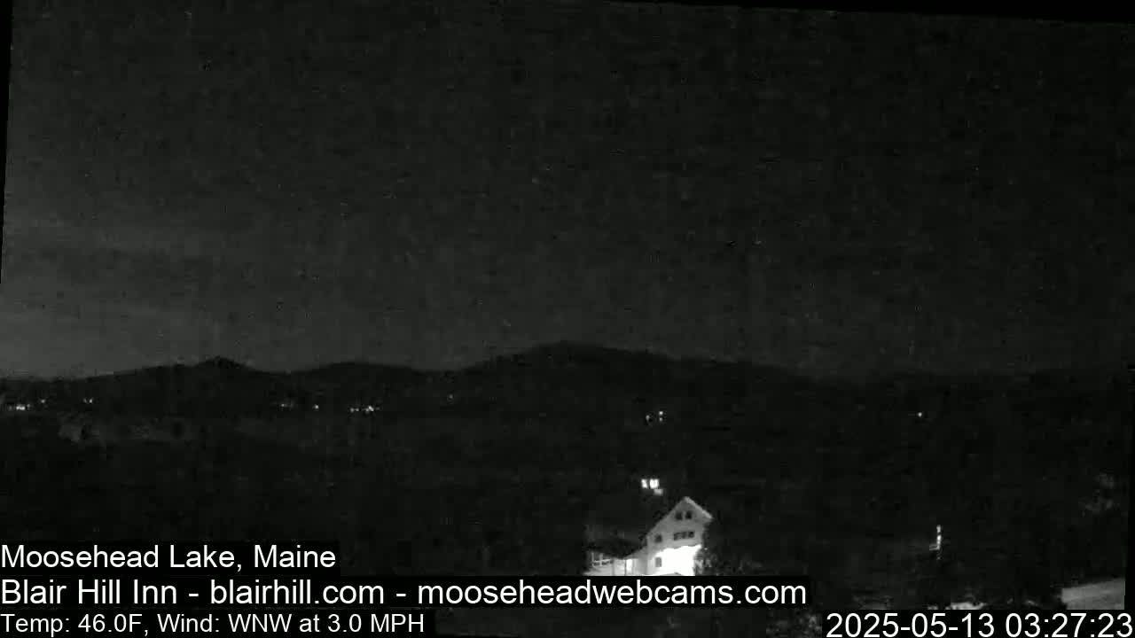 A nighttime view of a dark landscape with hills, a few distant lights, and a building, under a mostly clear sky with a 3 mph wind and a temperature of 46°F.