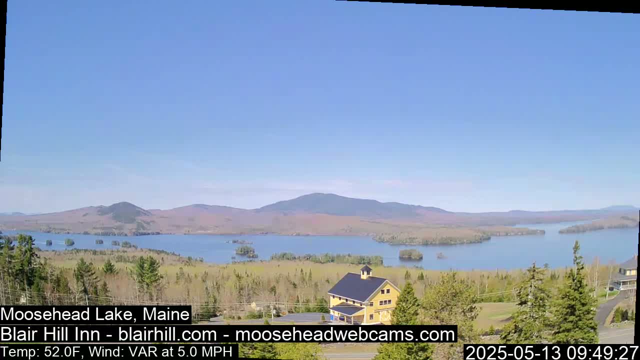 A large lake surrounded by trees and mountains is visible under a clear blue sky, with a yellow building in the foreground.
