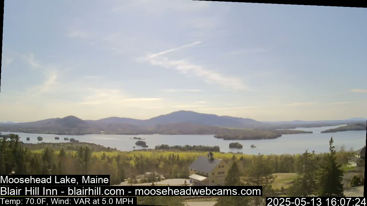 A picturesque view of a lake nestled amongst rolling hills and lush green trees under a mostly clear sky.