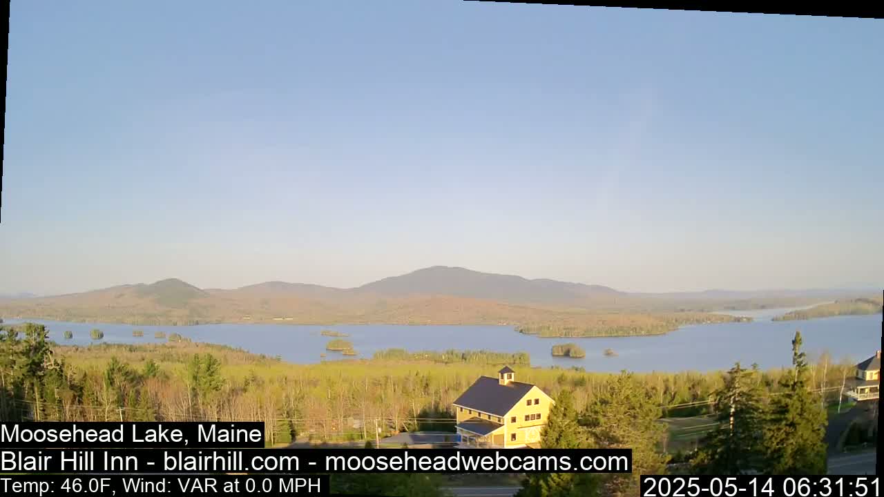 A calm, clear day reveals a view of a lake surrounded by trees and mountains, with a yellow building visible in the foreground.