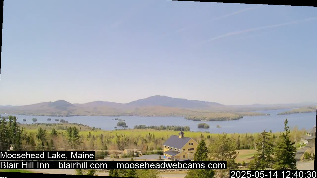 A large lake is visible under a mostly clear sky, surrounded by green trees and hills, with a yellow building in the foreground.