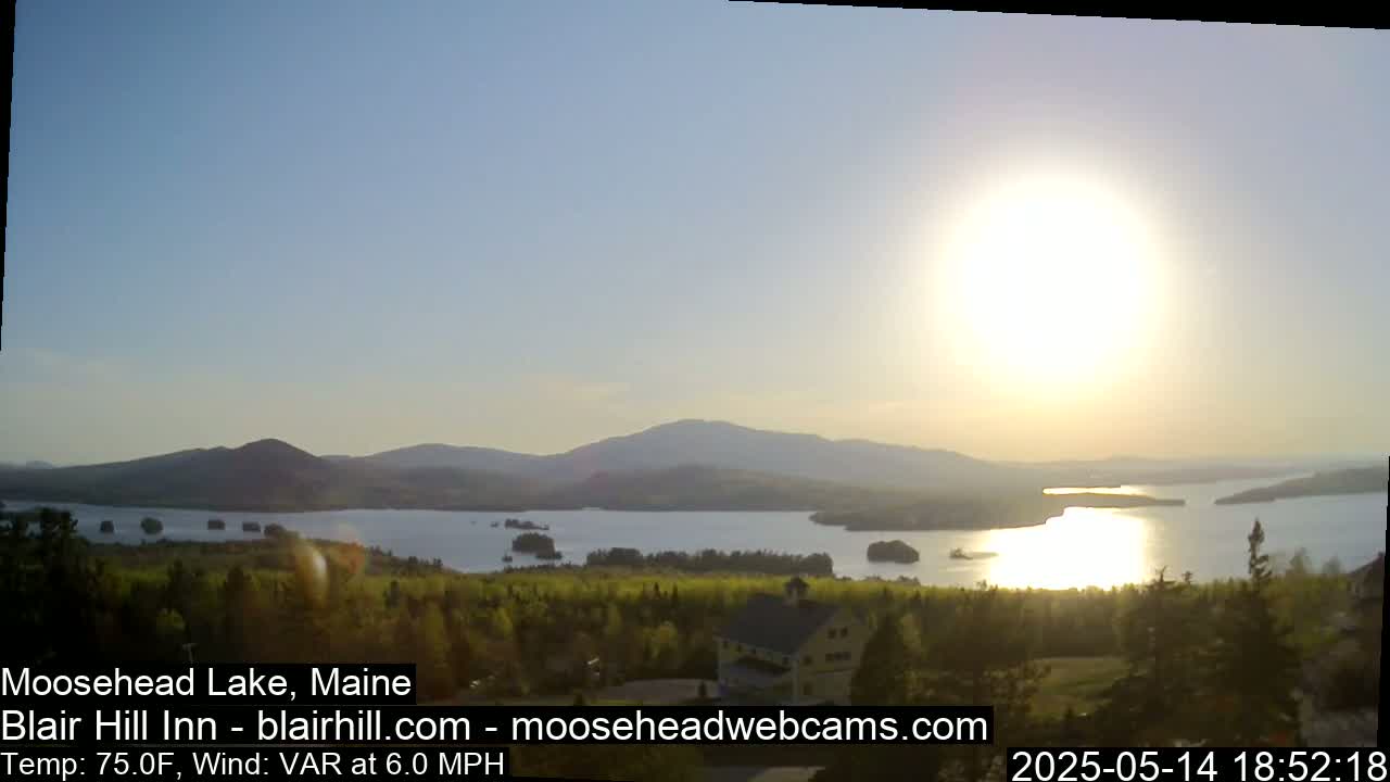 A calm lake reflecting the bright sun, surrounded by green trees and distant mountains under a clear sky.
