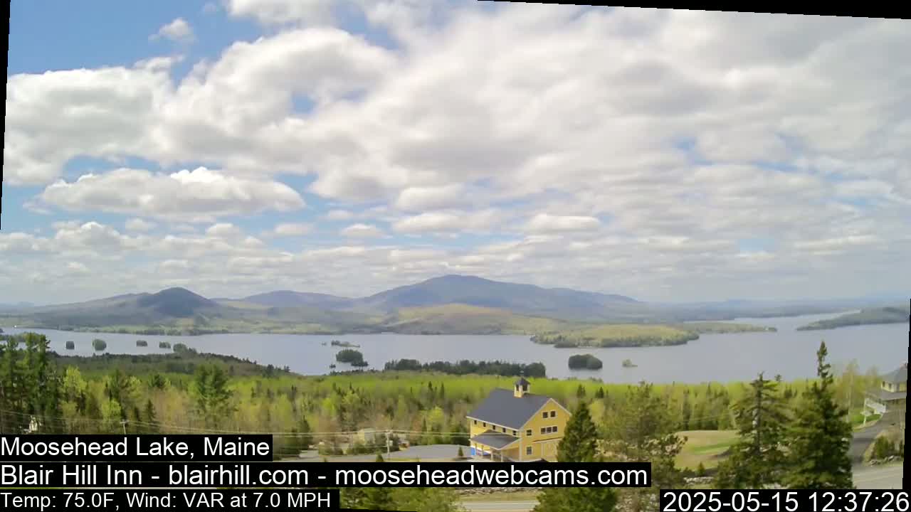 A partly cloudy day reveals a view of a large lake surrounded by green trees and distant mountains, with a yellow building visible in the foreground.