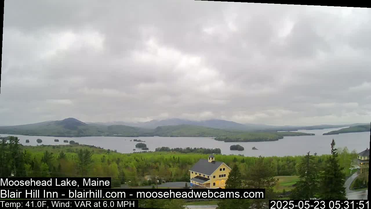 A large lake with islands and distant mountains is seen from a high vantage point under an overcast sky, a yellow building is visible in the foreground.